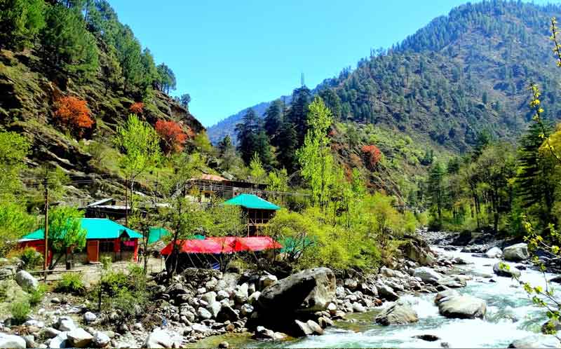 Houses around the river flowing and mountain ranges along with trees in Tirthan valley